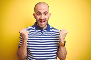 Naklejka premium Young bald man with beard wearing casual striped blue t-shirt over yellow isolated background celebrating surprised and amazed for success with arms raised and open eyes. Winner concept.