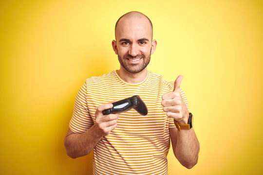 Young man playing video games using joystick gamepad over isolated yellow background happy with big smile doing ok sign, thumb up with fingers, excellent sign