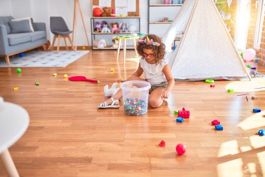 Beautiful toddler wearing glasses and unicorn diadem sitting on the floor playing with building blocks at kindergarten