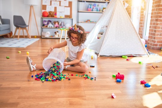 Beautiful toddler wearing glasses and unicorn diadem sitting on the floor playing with building blocks at kindergarten