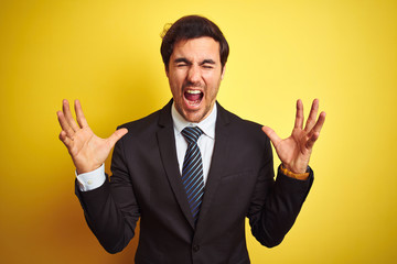 Young handsome businessman wearing suit and tie standing over isolated yellow background celebrating mad and crazy for success with arms raised and closed eyes screaming excited. Winner concept