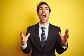 Young handsome businessman wearing suit and tie standing over isolated yellow background crazy and mad shouting and yelling with aggressive expression and arms raised. Frustration concept.