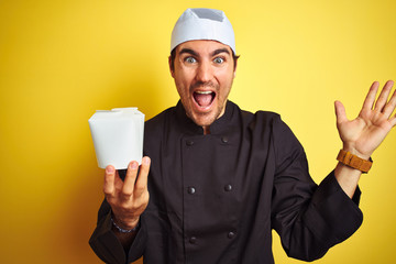 Young chef man wearing uniform and hat holding delivery box over isolated yellow background very...