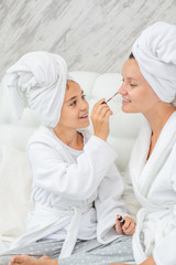 Obraz na płótnie Canvas Happy family at home. Mother and daughter are doing make up and having fun sitting on the bed at home. Mom and child girl are in bathrobes and with towels on their heads