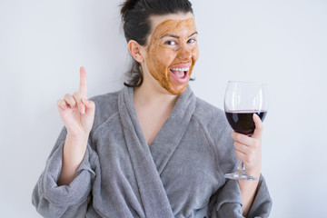 Beautiful woman wearing cosmetic facial mask as skincare treatment drinking glass of wine surprised with an idea or question pointing finger with happy face, number one