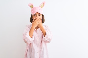 Beautiful child girl wearing sleep mask and pajama standing over isolated white background shocked covering mouth with hands for mistake. Secret concept.