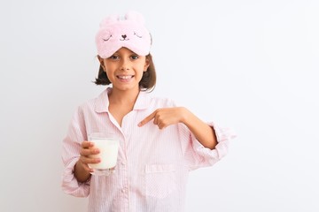 Child girl wearing sleep mask and pajama drinking glass of milk over isolated white background with surprise face pointing finger to himself