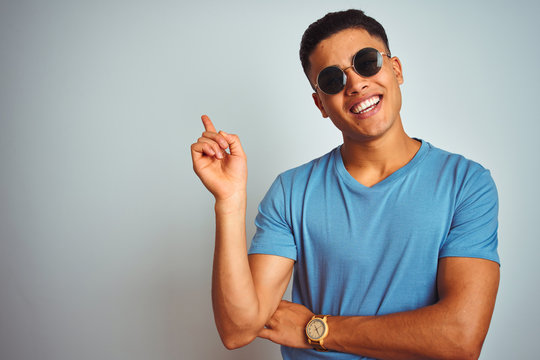 Young brazilian man wearing blue t-shirt and sunglasses over isolated white background with a big smile on face, pointing with hand and finger to the side looking at the camera.