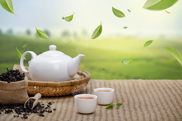 Warm cup of tea with teapot, flying green tea leaves in the air and dried herbs on the bamboo mat...