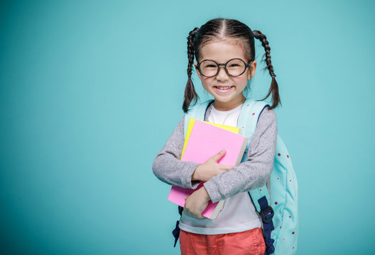 Beautiful smiling Asian little girl with glasses and hold a books with school bag is back to school, empty space in studio shot isolated on colorful blue background, Educational concept for school