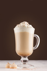Cappuccino coffee in elegant glass with whipped cream, cocoa powder and cookies on white table and dark brown wall, vertical.