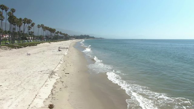 Cinematic horizontal moving aerial of beautiful empty beach with small waves and palm trees in Santa Barbara, California, USA. It's a sunny and warm afternoon on this public beach.