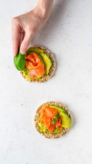 Hand laying a leaf of basil on open face sandwiches. Buckwheat crispbread toast with avocado, salmon, flax seeds.