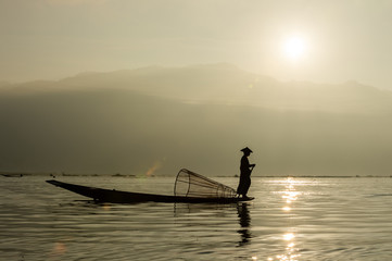 Silhouette Burmese fisherman wearing a hat standing at the back of a boat rowing with legs in the morning, mist faded at Inle Lake, Shan State in Myanmar.