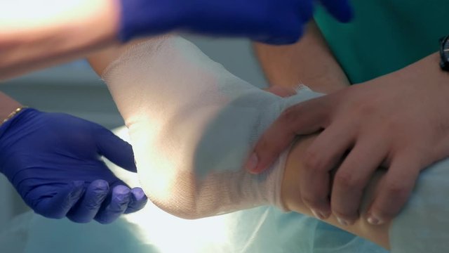 Nurse and doctor surgeon bandage the patient's leg foot after surgery to ankle hygroma removal, closeup view. Operation in operating room in hospital. One day surgery concept.