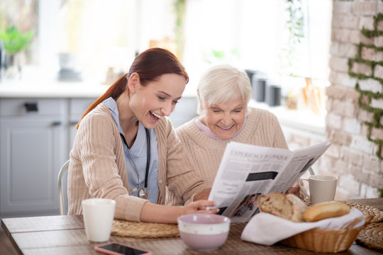 Retired woman and caregiver laughing while reading newspaper