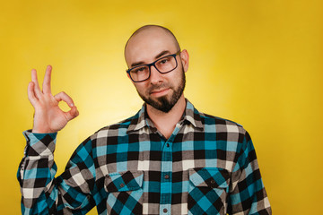 Naklejka premium People and emotions. A bald, bearded man in glasses and a blue checked shirt, fingers pointing to the ok sign. Yellow background. Copy space