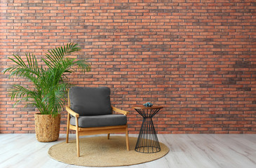 Modern room interior with stylish grey armchair and potted plant near brick wall