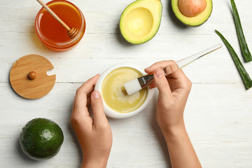 Woman with handmade face mask, brush and ingredients at white wooden table, top view