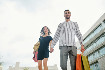 Happy excited young man holding hand of his girlfriend when walking to another store after shopping in mall