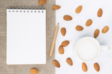 Fototapeta na wymiar Almond milk in cup, notebook, pencil, nuts on white concrete background. Non-dairy, vegan, recipe concept. Top view, flat lay, copy space, mock up