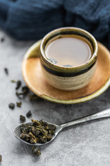 A cup of hot green tea with dry leaves in spoon on cement table. Healthy drink concept.