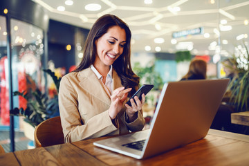 Dedicated attractive caucasian businesswoman dressed in beige jacket using smart phone while sitting in cafe. On table is laptop.