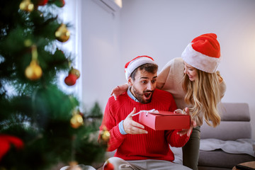 Charming caucasian woman giving christmas gift to her boyfriend. Both are dressed in sweaters and having santa hats on heads. Living room interior, christmas holiday concept.