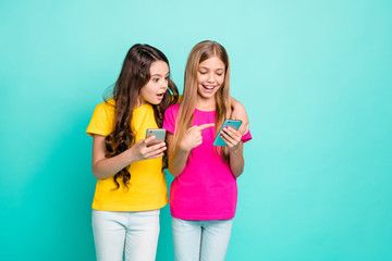 Photo of funny casual nice cute friendly couple of blonde brunette girlfriends wearing pink yellow t-shirts getting shocked with something oh their phones while isolated with teal background