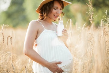 Beautiful young pregnant woman in a wheat field drinks milk from a glass bottle. Portrait of a...