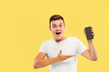 Caucasian young man's half-length portrait on yellow studio background. Beautiful male model in shirt. Concept of human emotions, facial expression, sales, ad. Showing phone's screen and smiling.