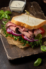 Roast beef sandwich with tomato, onion, lettuce and mustard sauce on dark background. Delicious...