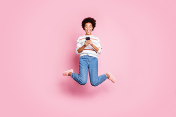 Full length body size photo of cheerful fascinating rejoicing cute girl browsing through her telephone jumping wearing jeans denim striped sweater isolated over pastel color background