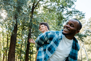 Fototapeta premium low angle view of happy african american piggybacking cute curly son near soap bubbles in park