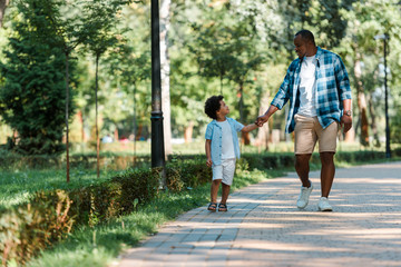 happy african american kid holding hands with handsome father while walking in park