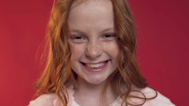 Beautiful picture of charming red headed teen girl with long hair and freckles looking at camera with nice smile on the red background