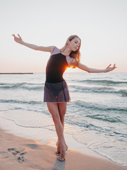 Fototapeta premium Dancing ballerina in black silk dress on embankment near ocean or sea at sunrise or sunset. Silhouette of young woman with long hair practicing classic exercises with emotions