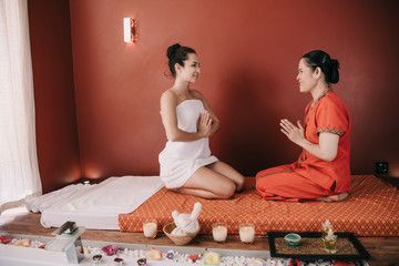 smiling woman and asian masseur sitting on massage mat and showing greeting hands