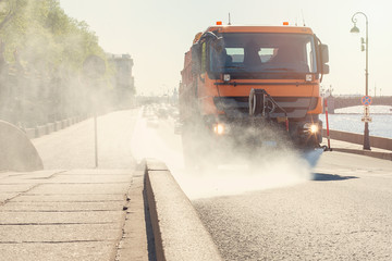 Watering machine washes city road with water. Big orange watering machine washes the road part with water. Sun day in Saint-Petersburg, Russia