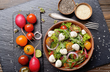 Flat lay of vegetable salad with mozzarella cheese, lettuce, cherry tomatoes, radish, cucumber, onion and basil in clay dish