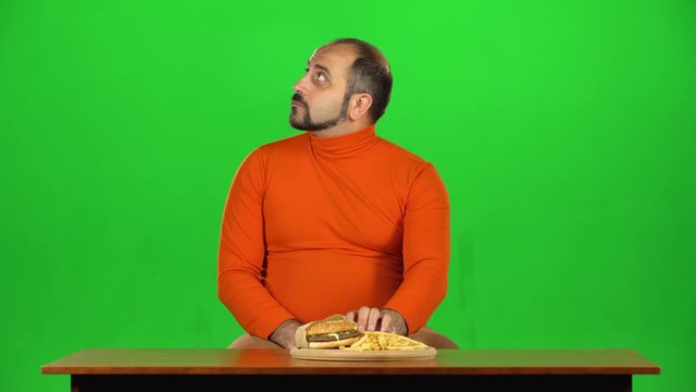 Man with overweight looks at delicious junk foods on the table and resisting unhealthy eating, green screen