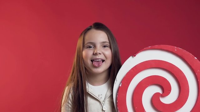 Close up of funny smiling teen girl with braces showing tongue,holding plastic lollipop in hand on the red background