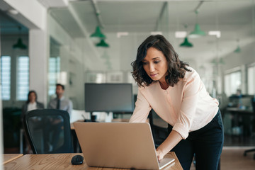 Businesswoman leaning over her office desk working on a laptop