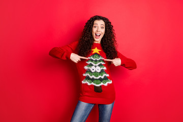 Portrait of amazed funny funky crazy girl point at her seasonal comfort jumper pullover recommend her sweater with christmas tree decor for theme x-mas noel party isolated over red color background