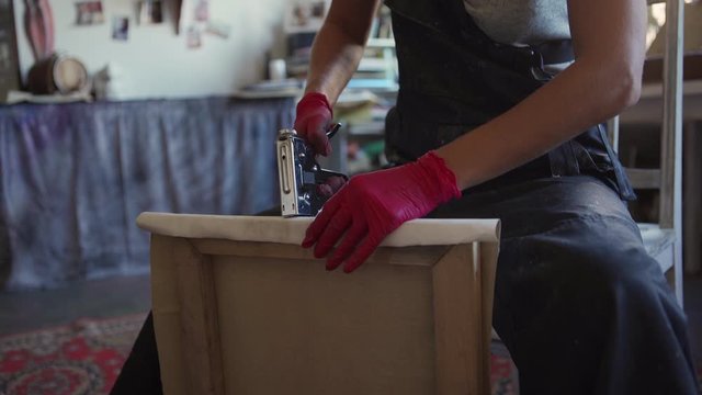 Unrecognizable female painter using staple gun to stretch canvas on frame sitting on chair in art studio