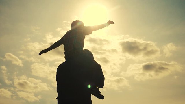 happy family silhouette teamwork concept slow motion video. son boy superhero sitting on his father man neck lifestyle depicts a flight of an airplane playing a pilot. father and son silhouette at