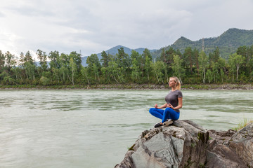 Blond beautiful woman sitting on the stone cliff ground resting and doing yoga and meditation outdoors in the mountains near the river. Workout and relaxation in nature.