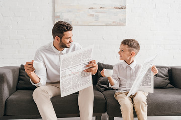 son and dad in formal wear sitting on sofa and reading newspapers