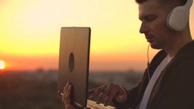A man freelancer in headphones standing on the roof at sunset writes on the keyboard code pages. Little business. Listen to music and work at the computer enjoying the beautiful view from the roof