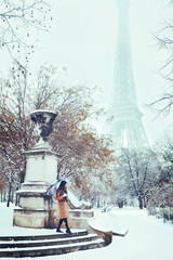 A young woman in a beige coat walks under an umbrella in a snowy winter Paris against the backdrop of the Eiffel Tower. Romantic brunette girl is walking in the background of falling snow in Paris. 